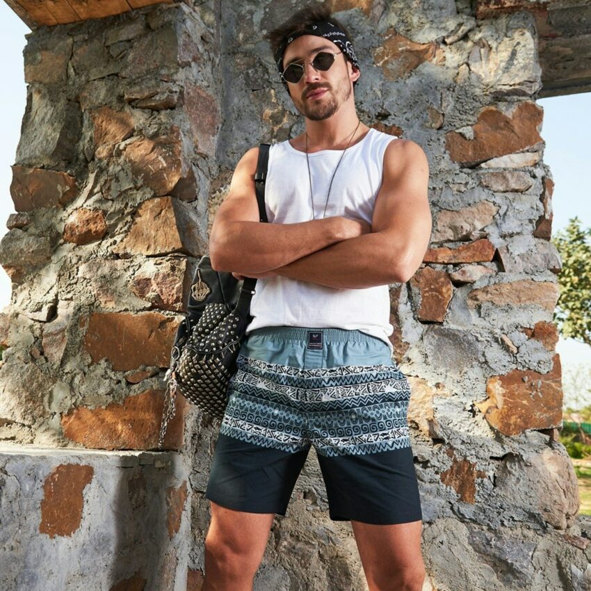 man in white tank top and black and white plaid shorts standing beside brown rock
